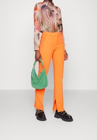 Pantalon orange sur mesure avec fentes latérales, chemise longue à manches longues à fleurs multicolores, et sac à main en daim vert. Bottes noires épaisses avec des accents dorés.