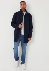 Marineblauer Mantel mit hohem Kragen und Druckknopfverschluss, aus weichem Material gefertigt. Kombiniert mit hellblauen Jeans und weißen Sneakers.