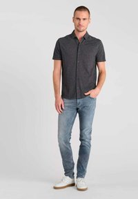 Korte mouwen, knoopsluiting overhemd in donkergrijze stof, gecombineerd met lichtblauwe jeans en witte sneakers. Model staat tegen een neutrale achtergrond.