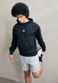 Črna kapučna majica s sprednjim žepom in logotipom, v kombinaciji z sivimi kratkimi hlačami. Model raztegne nogo, kar prikazuje športno obleko v minimalističnem okolju.