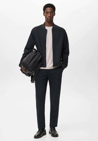 Chaqueta bomber negra sobre una camisa de color beige claro, combinada con pantalones oscuros y zapatos negros, sosteniendo una mochila negra. Detalles de diseño minimalista.