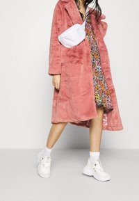 Nepvacht, lange roze jas met revers, gelaagd over een multicolore bloemenjurk. Een witte crossbody tas en chunky witte sneakers maken de look compleet.