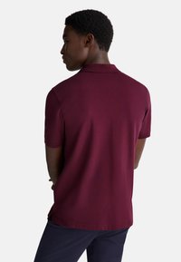 Giovane visto da dietro che indossa una polo bordeaux a maniche corte e pantaloni scuri su uno sfondo chiaro e semplice.