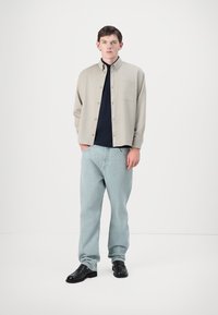 Camicia button-up grigia chiara sopra un maglione blu navy, abbinata a jeans blu chiaro e scarpe nere. L'outfit presenta un dettaglio con taschino sul petto.