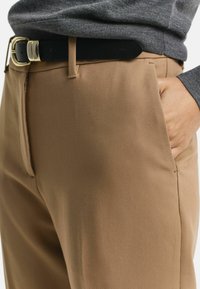 Tan pants with a straight cut, lightweight material, side pockets, and a black belt featuring a gold buckle. Gray sweater peeking at the waist.
