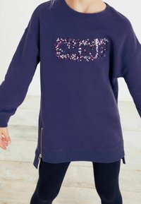 Fialová mikina s lesklým flitrovým nápisem "SHINE", dlouhé rukávy, boční zipový detail a žebrované manžety. V kombinaci s černými legínami.