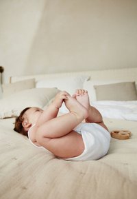 Bebeluș în combinezon alb întins pe o pătură bej texturată, întinzând mâna către degetele de la picioare, cu o jucărie de lemn alături, așternut moale în fundal.