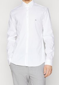 Man i en vit långärmad skjorta med knappar och en liten "CK"-logotyp på bröstet, kombinerad med ljusgrå byxor.