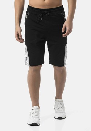 Man draagt zwarte shorts met trekkoorden en witte zijstrepen en witte sneakers, staand tegen een effen achtergrond.