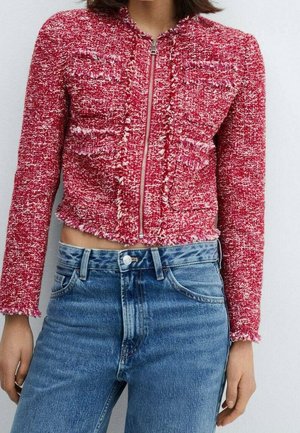 Femme portant une veste rouge texturée à fermeture éclair courte avec bords frangés, associée à un jean en denim bleu taille haute.