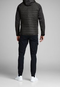 Giacca nera con texture trapuntata sulla schiena, abbinata a pantaloni cargo scuri e sneakers bianche. Presenta un cappuccio e polsini elasticizzati.