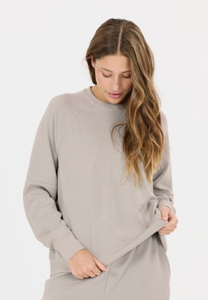 Kvinde med langt, bølget hår iført en løs beige sweatshirt og matchende bukser, der kigger ned og holder i kanten af sweatshirten.