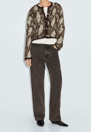 Personne portant un cardigan boutonné marron et beige à motif flou, un t-shirt blanc, un jean ample foncé et des chaussures noires pointues.