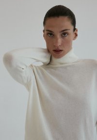 Kremowy sweter z golfem o miękkiej fakturze, luźnym kroju i żebrowanym wykończeniu przy kołnierzu. Modelka pozuje z jedną ręką na szyi.