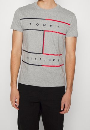 Mężczyzna w szarej koszulce Tommy Hilfiger z czerwono-czarną prostokątną grafiką i czarnych spodniach.