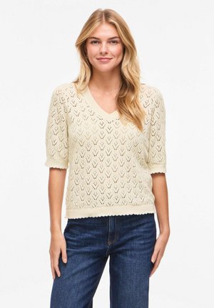 Jauna sieviete ar gariem blondiem matiem, ģērbusies krēmkrāsas īss piedurkņu adītā džemperī un zilās džinsās, stāvot un smaidot baltā fonā.