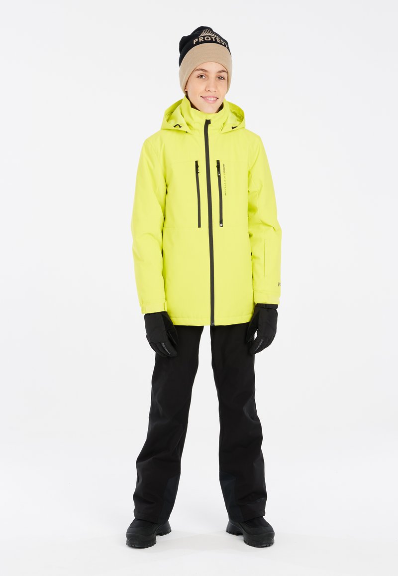 Veste de ski jaune vif avec des poches zippées noires, portée sur un pantalon noir et des gants. Le col est haut et il y a une capuche attachée.