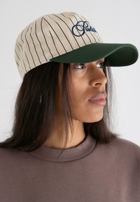 Casquette beige avec des rayures verticales bleu marine et un bord vert, ornée d'un texte brodé en bleu. Tissu lisse avec une conception structurée.