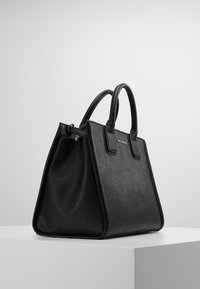 Musta, rakenteellinen käsilaukku, joka on valmistettu karheatuntuisesta synteettisestä materiaalista. Laukussa on kaksi yläkahvaa, kultainen vetoketjukiinnitys ja logoelementti edessä.