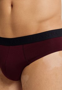 Bordeauxfarbene Herrenslip mit einem schwarzen elastischen Bund, der mit eingeprägten Logodetails versehen ist. Glattes Material mit passgenauer Form und Nahtakzenten.