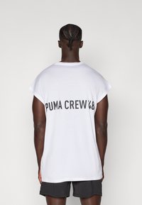 T-shirt blanc à manches courtes avec le texte noir "PUMA CREW '48" au dos. Le tissu semble doux avec une légère texture. Associé à un short noir.