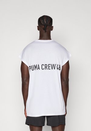 Hvid T-shirt med korte ærmer, der har sorte bogstaver "PUMA CREW '48" på ryggen. Stoffet ser blødt ud med en let tekstur. Kombineret med mørke shorts.