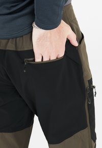 Hånd med sølvarmbånd delvist inde i lynlåslukket sidelomme på sorte og olivengrønne cargobukser, båret med mørk langærmet top.