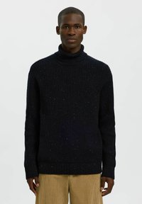 Jersey de cuello alto de punto azul marino con una textura de canalé y un sutil patrón moteado, combinado con pantalones de pana color beige claro.