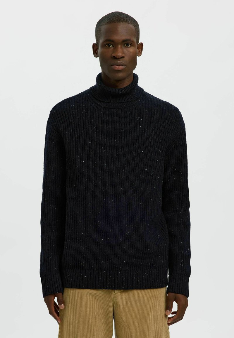 Jersey de cuello alto de punto azul marino con una textura de canalé y un sutil patrón moteado, combinado con pantalones de pana color beige claro.