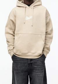 Beige Kapuzenpulli aus weichem Material, mit einer Fronttasche, Kordeln und einem weißen Logo auf der Brust. Getragen mit dunklen Jeans.