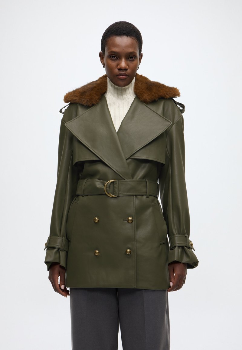 Trench-coat en cuir vert olive avec col en fausse fourrure marron, taille cintrée par une ceinture, boutons dorés en accent et revers structurés. Le mannequin porte un pull crème.
