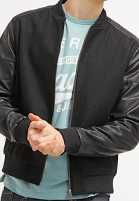Černá bomber bunda s koženými rukávy, žebrovanými manžetami a pasem, se zapínáním na zip vpředu a designem s kulatým výstřihem.