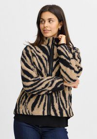 Veste en polaire rayée beige et noire avec un col montant et une fermeture éclair complète, dotée d'un tissu doux et texturé ainsi que de poignets élastiques.