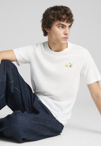 Camiseta blanca de manga corta hecha de algodón, con un logo de Puma multicolor en el pecho. Combinada con pantalones de mezclilla oscuros.