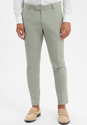 Man draagt lichtgroene, nauwsluitende pantalon, gecombineerd met beige loafers en een wit overhemd met lange mouwen, staand tegen een witte achtergrond.