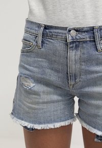 Pantalones cortos de mezclilla en azul claro con dobladillo deshilachado y detalles desgastados. Cuentan con cinco bolsillos y cierre de botón metálico en la cintura.