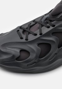 Svarta sneakers med en texturerad yta, framträdande utskärningar och platta snören. Materialet verkar syntetiskt med en strömlinjeformad design.