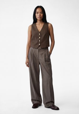 Nainen seisoo käsineen taskuissa, yllään ruskea hihaton napitettava liivi, harmaat leveälahkeiset housut ja tummanruskeat loafer-kengät valkoisella taustalla.