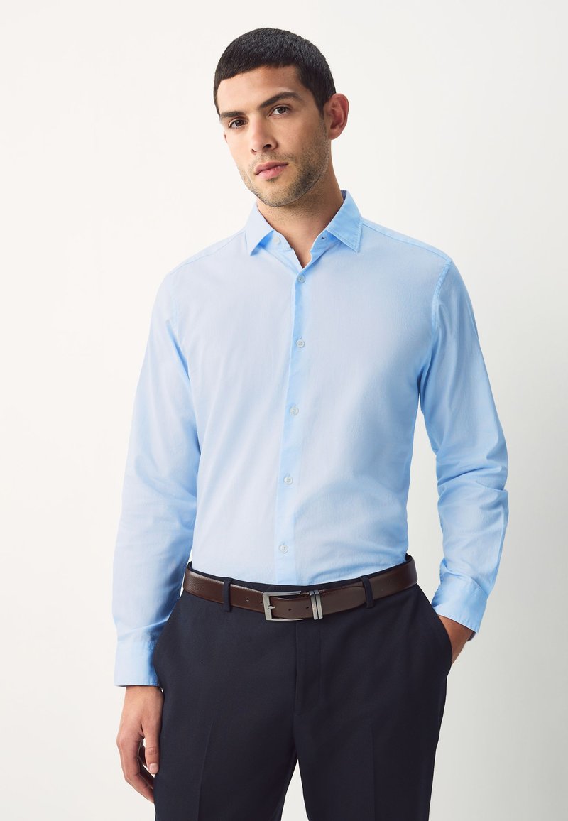 Chemise bleu clair à manches longues en tissu de coton, avec un col pointu et une fermeture à boutons, associée à un pantalon foncé et une ceinture marron.