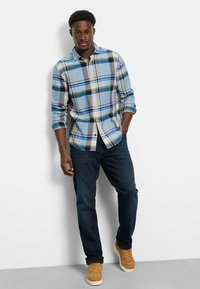 Blau kariertes Button-Down-Hemd mit unterschiedlich gefärbten Streifen, langen Ärmeln und einem lässigen Schnitt, kombiniert mit dunkelblauen Jeans und hellbraunen Sneakers.