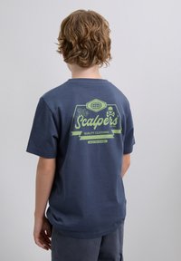 Junge mit welligem blonden Haar steht mit dem Rücken zur Kamera und trägt ein marineblaues T-Shirt mit grünem Text und einem Totenkopf-Logo auf dem Rücken.