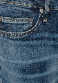 Gros plan d'un jean en denim bleu montrant la poche avant, la boucle de ceinture et les détails de surpiqûres orange sur un tissu texturé.