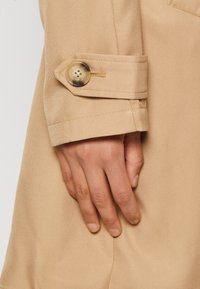 Beige ballonkabát mandzsetta, amelyen egy nagy, kerek gomb található négy lyukkal, sima szövettextúrával és precíz varrási részletekkel.