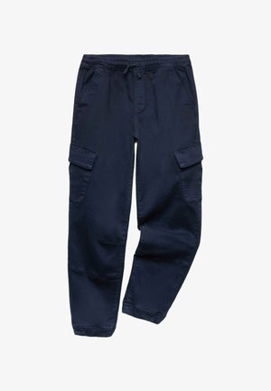 Dreng står med armene krydsede iført en brun og lyseblå striped langærmet skjorte, mørke cargobukser og hvide sneakers på hvid baggrund.