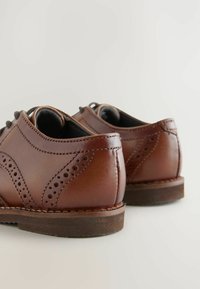 Brązowe skórzane buty z detalami w kształcie skrzydeł, okrągłym noskiem i niskim obcasem. Posiadają perforowane akcenty oraz kontrastowe szwy wzdłuż szwów.