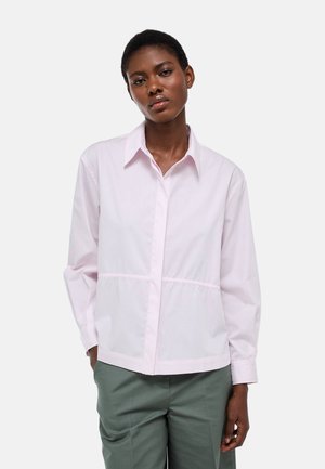 Femme portant une chemise rose pâle à boutons avec col et manches longues, associée à un pantalon taille haute gris-vert.