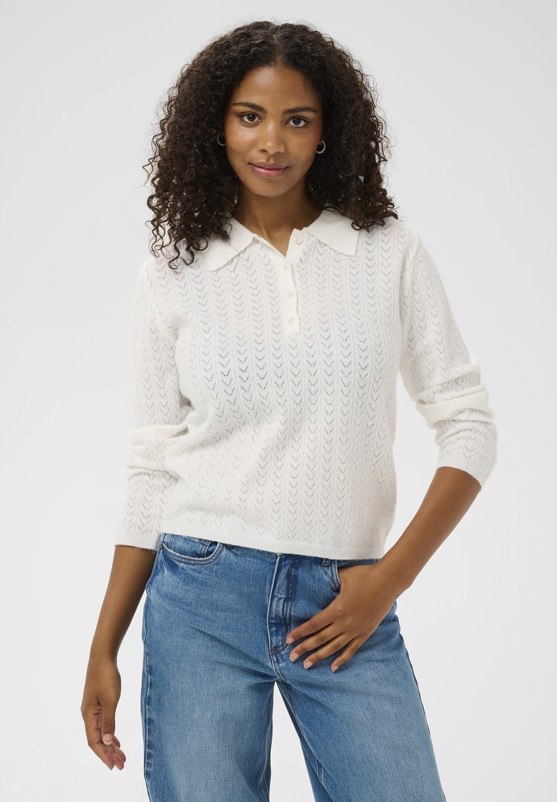 Pull en tricot blanc avec un col, trois boutons et un motif en chevron texturé. Associé à un jean en denim bleu.