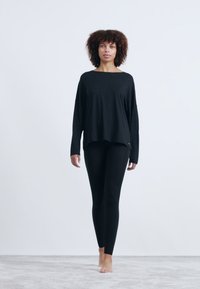 Camisola preta de mangas longas com um padrão texturizado e ajuste solto, combinada com leggings pretas. O modelo está descalço, em pé sobre uma superfície macia.