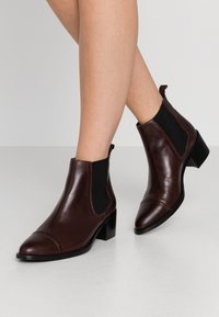 Botine din piele maro, cu bot ascuțit, panouri laterale din elastic și toc mic, având o textură netedă și un design minimalist.