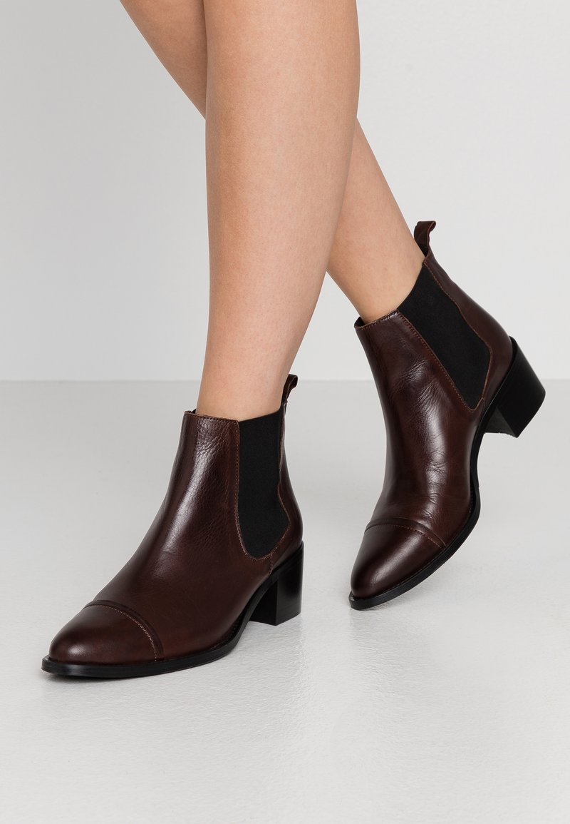 Botine din piele maro, cu bot ascuțit, panouri laterale din elastic și toc mic, având o textură netedă și un design minimalist.